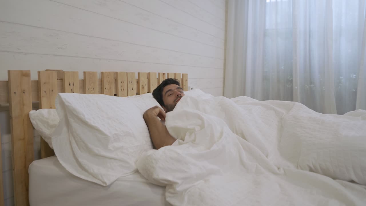 Giovane uomo mediorientale assonnato che si sveglia. Persone sdraiate e che dormono sul letto in camera da letto la mattina presto a casa. Stile di vita