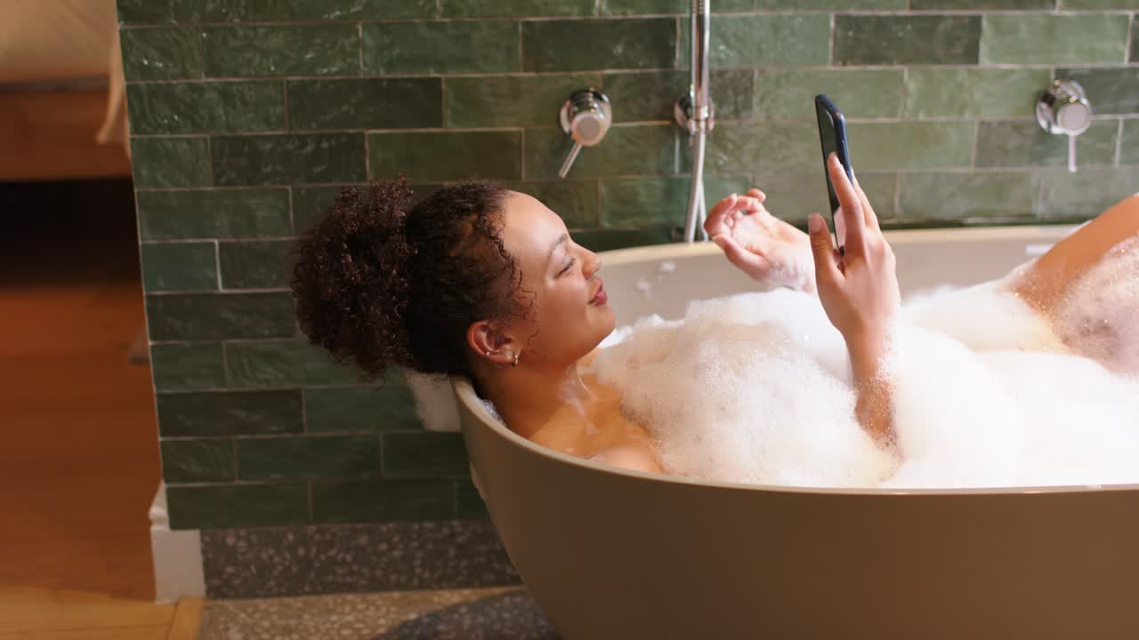 African American woman 20s reclining in bathtub with bubbles lifting phone and reacting to screen