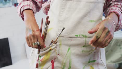 sección media de artista femenina biracial poniendo pinceles de pintura en el bolsillo del delantal en el estudio, cámara lenta