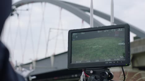 Handheld-camera-perspective-of-a-white-drone-with-four-rotors,-flying-near-a-modern-cable-arch-bridge,-with-overcast-gloomy-sky-backdrop-from-monitor-perspective