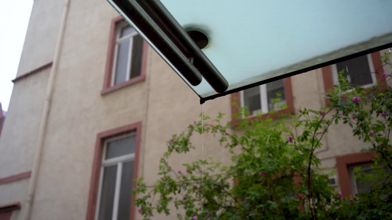 Raindrops falling from a building's eave against a blurred residential backdrop, focus on water droplet