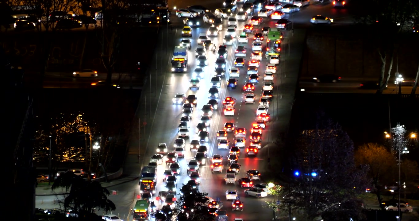 El tráfico intenso llena varios carriles de una carretera de Tiflis por la noche, con brillantes faros blancos y luces traseras rojas en corrientes opuestas vistas desde un punto elevado