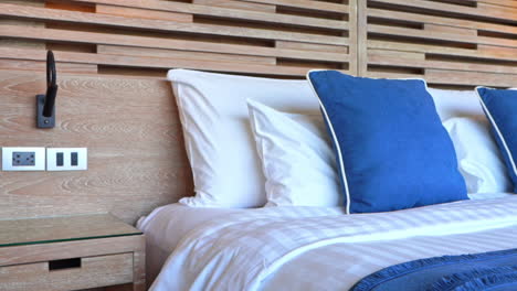 hotel room interior with king's size made-up bed, white and blue pillow and wooden headboard daytime