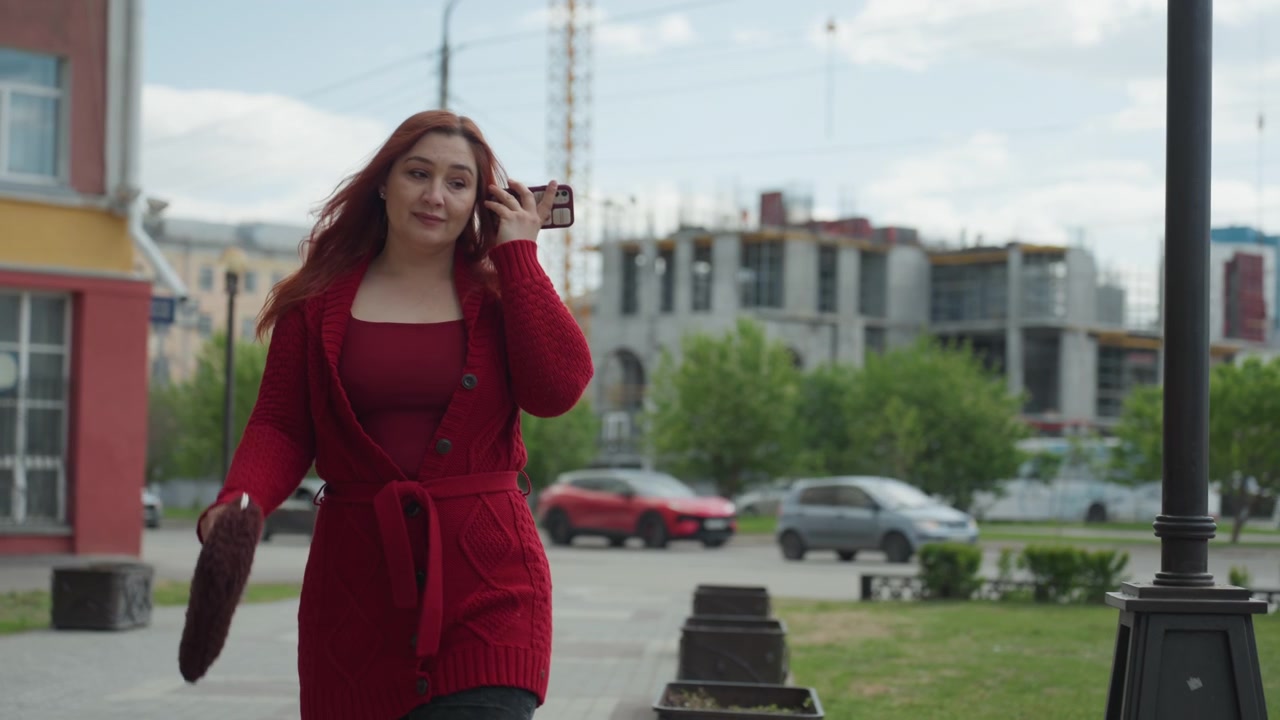 Close up of woman walking outdoors while talking on her phone and carrying items. Woman wears red clothing holding one object in each hand as woman moves through urban environment