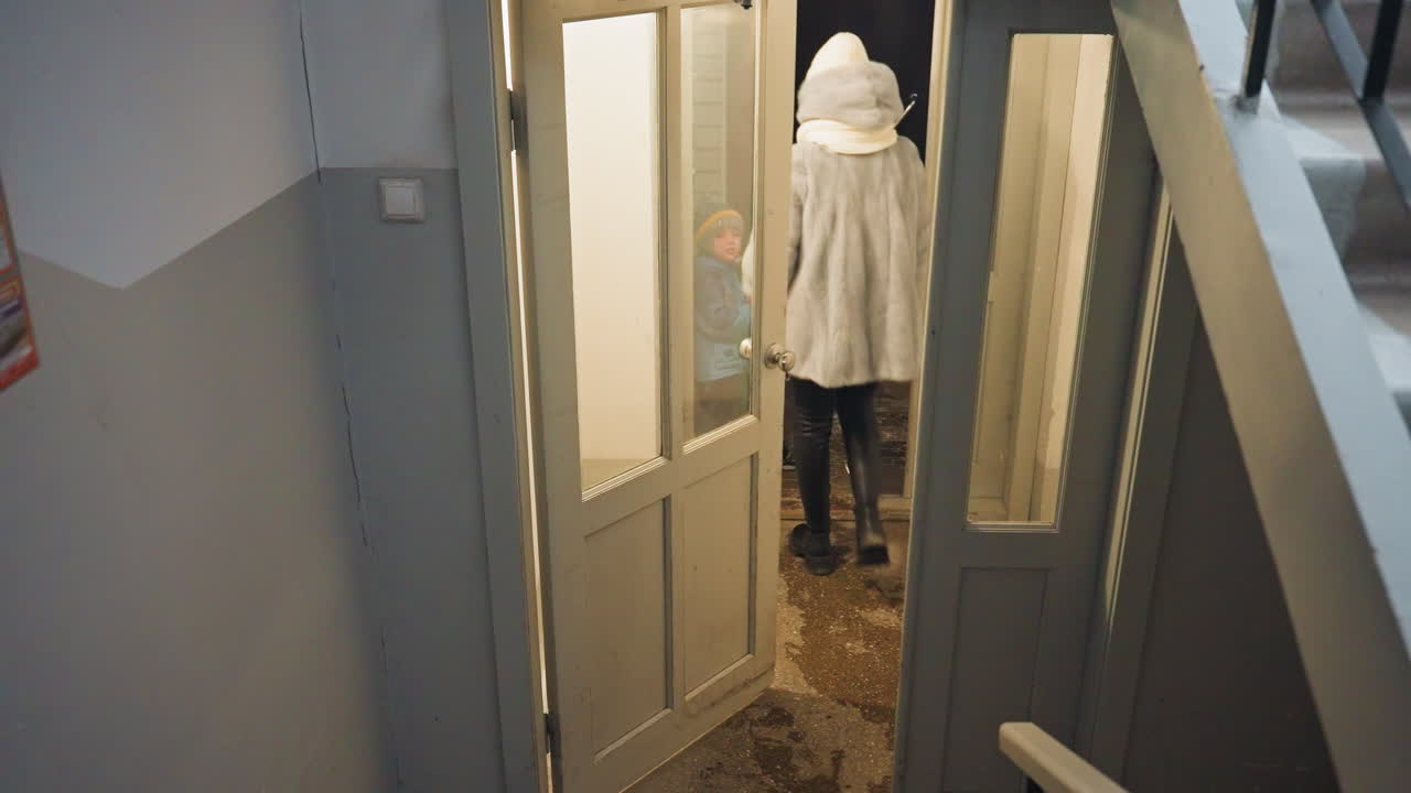 Mamma och son i dörröppningen, mamman i vinterrock går in i trapphuset och öppnar glasdörren medan den lille sonen tittar inifrån genom fönstret, varmt lampglitter kastar mjukt sken, tyst återförening, skyddande