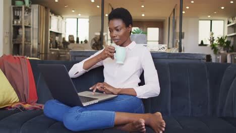 Happy-african-american-woman-sitting-on-sofa-in-living-room,-using-laptop