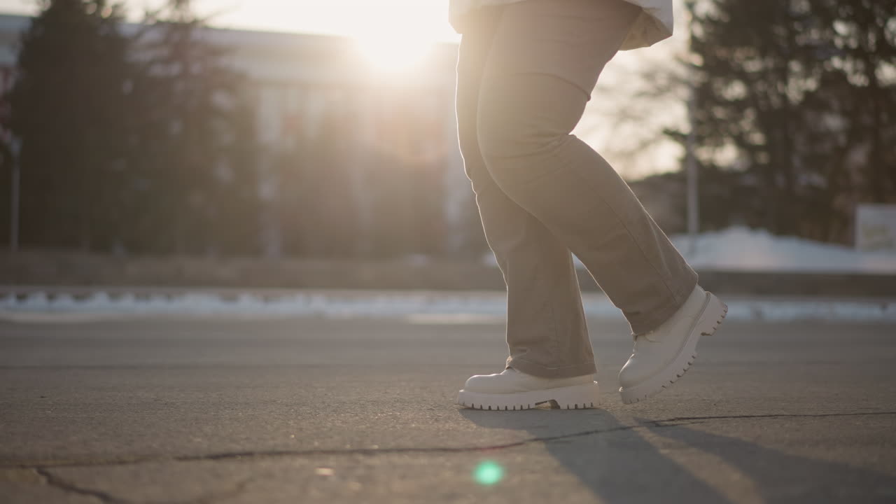 Young woman moving with rhythm across sunlit pavement in white boots and winter pants, foot taps creating energetic winter beat on urban asphalt stage under warm glowing sunset light