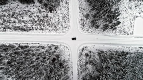 Vista-Aérea-De-Pájaro-De-Un-Coche-Negro-Parado-En-Medio-De-La-Encrucijada-Con-Nieve-Cubierta-En-Las-Carreteras-Y-Bosque-De-Pinos-En-Los-Cuatro-Lados