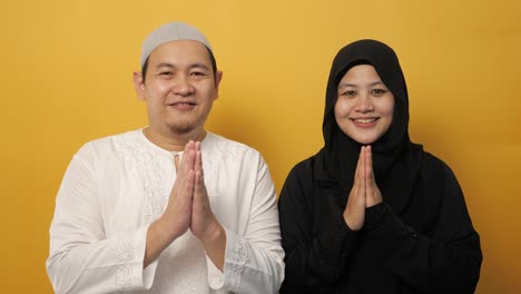 happy asian muslim couple making greeting gesture, hands raised up, islamic culture, against yellow