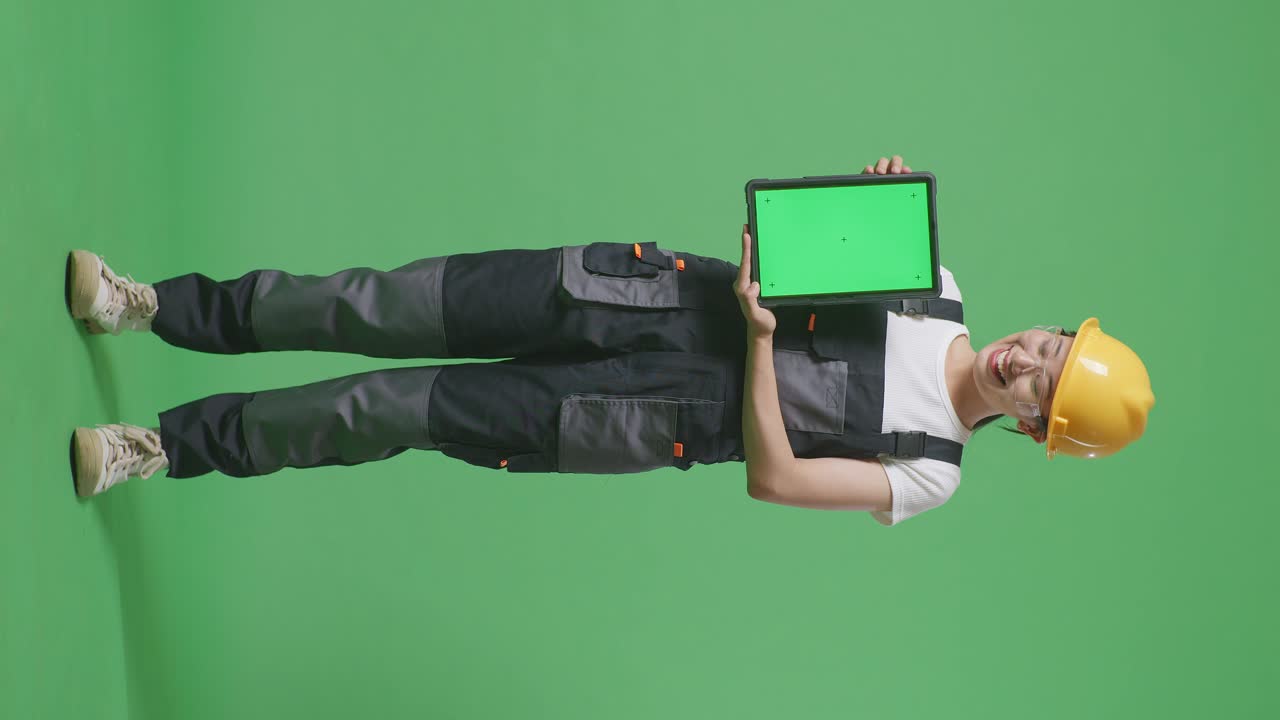 cuerpo lleno de mujer asiática trabajadora con gafas de protección y casco de seguridad sonriendo y mostrando la tableta de pantalla verde a la cámara mientras está de pie en el estudio de fondo de pantalla verde