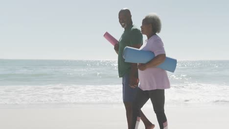 Glückliches-älteres-Afroamerikanisches-Paar,-Das-In-Zeitlupe-Am-Strand-Spaziert-Und-Yogamatten-Hält