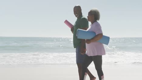 feliz pareja afroamericana de alto nivel caminando y sosteniendo esteras de yoga en la playa, en cámara lenta