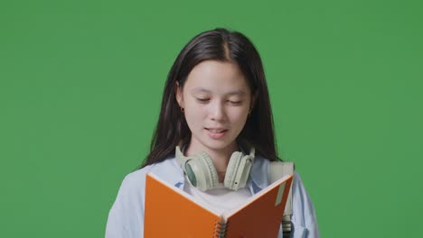 close up of asian teen girl student with a backpack holding and reading a book while standing in the green screen background studio