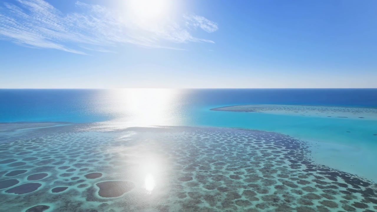 透明なターコイズ色のラグーンの空撮。サンゴ礁の模様が広がり、水面には強い太陽の反射が見られ、澄み切った青空の下に小さな島が浮かんでいます。