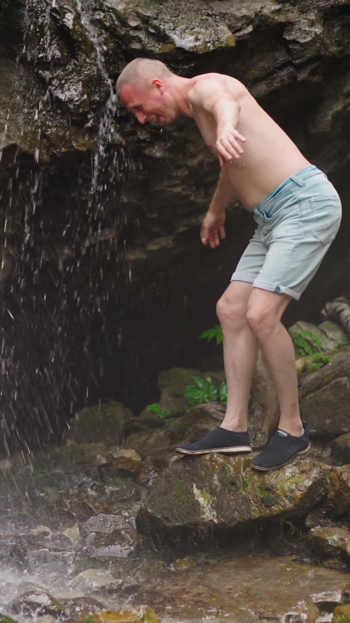Joyful man tourist wets bald head with clear waterfall streams standing on old slippy rock at virgin highland reserve on summer day slow motion