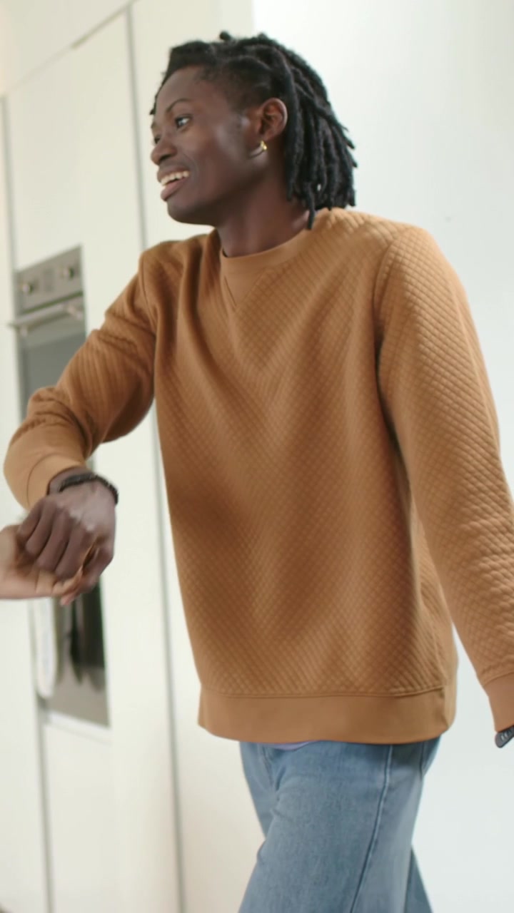 Vertical video: Smiling man replying offscreen, gesturing in kitchen near oven in brown sweater