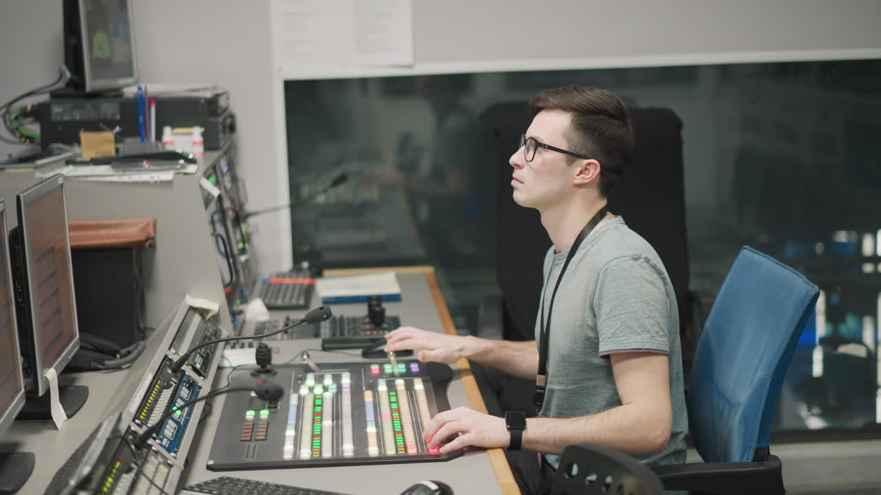 Man working in a studio, adjusting video production equipment, kneeling beside a monitor. Studio setup with bright lighting, reflective floors, and technical equipment in the background