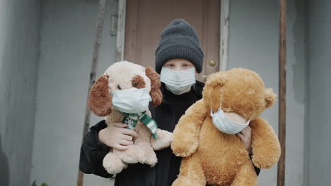 a sad child stands on the threshold of the house, holding two toys in protective masks. protect children concept