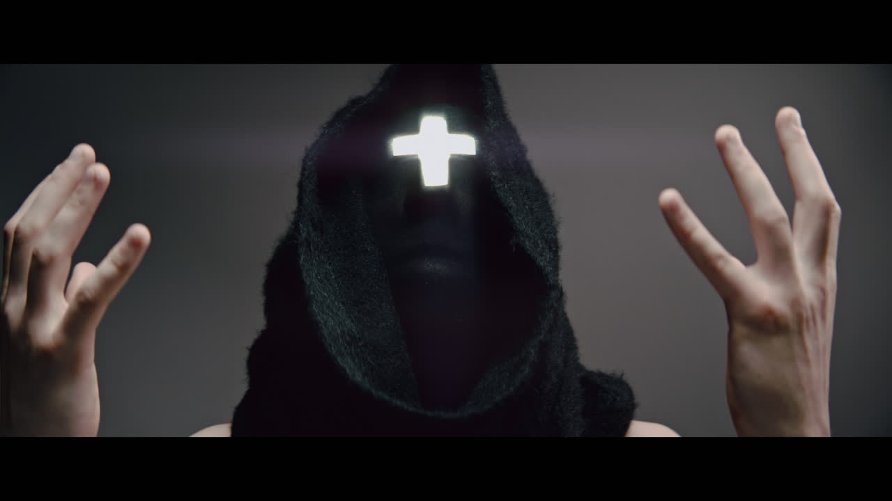Masked and hooded man with cross of light glowing on his face looks in the camera