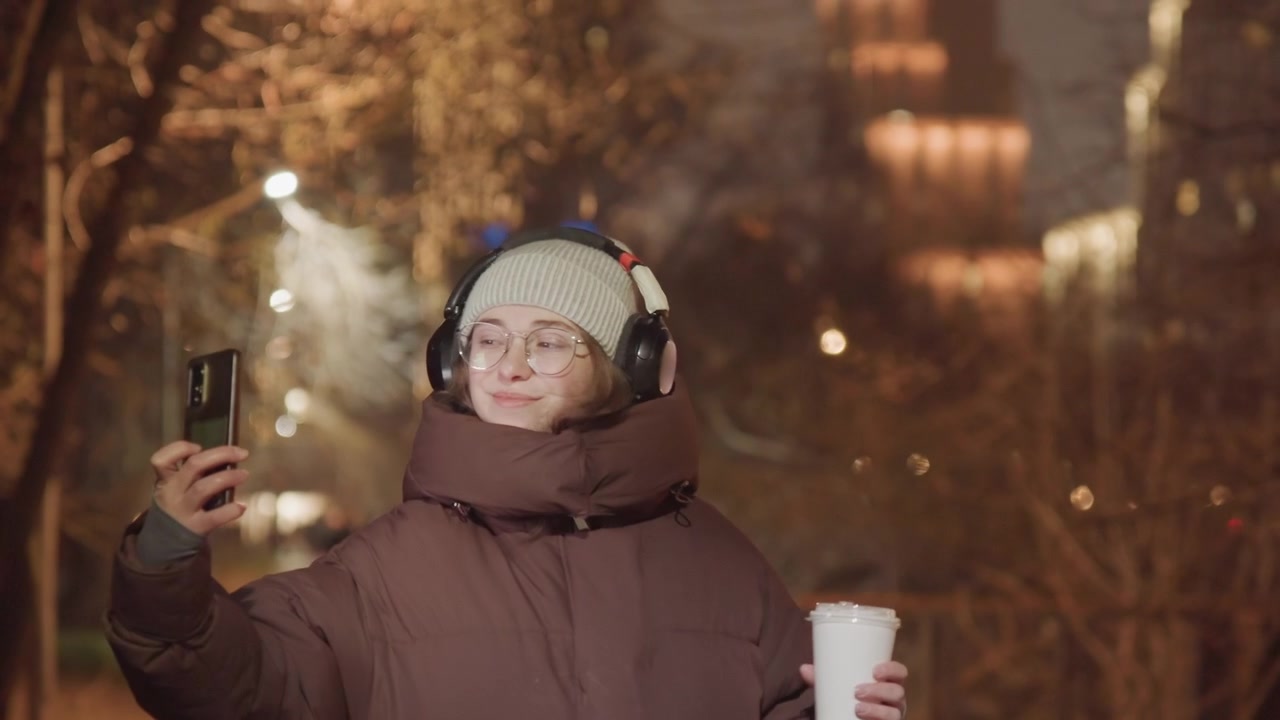 Giovane donna che si scatta un selfie di notte su un sentiero del parco con caffè in mano e cuffie, espressioni giocose e bokeh delle luci della città suggeriscono un'atmosfera da content creator, moda casual e passeggiata rilassata