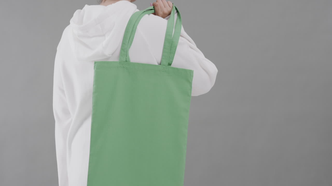 Donna caucasica che indossa una felpa bianca con cappuccio, tiene una borsa verde su sfondo grigio, spazio per il testo, movimento rallentato