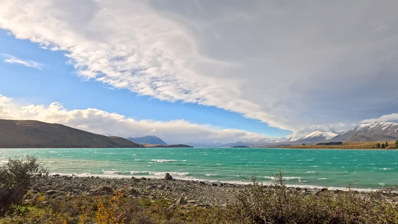 Turquoise lake with choppy waves, thorny shrubs in foreground, snow-capped mountains, dramatic clouds, and strong wind under bright daylight. Static wide shot