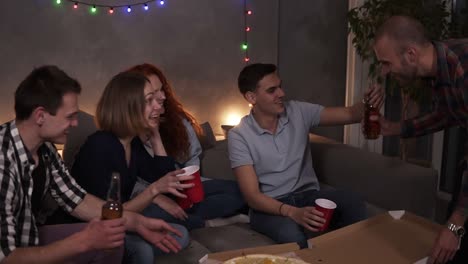 Cheerful-friends-got-delivery-pizza-and-celebrating-party-clinking-bottles-and-red-cups-with-beer-and-soda-sitting-on-sofa-at-loft-room.-So-excited-about-big-pizza-party.-Slow-motion