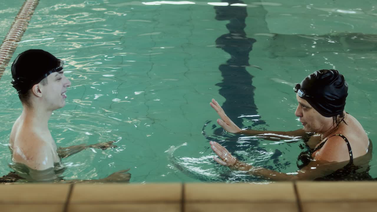 Two people in swim caps conversing in an indoor swimming pool