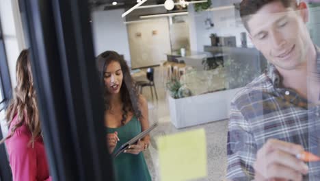 diverse creative colleagues in discussion making notes on glass wall in office, slow motion