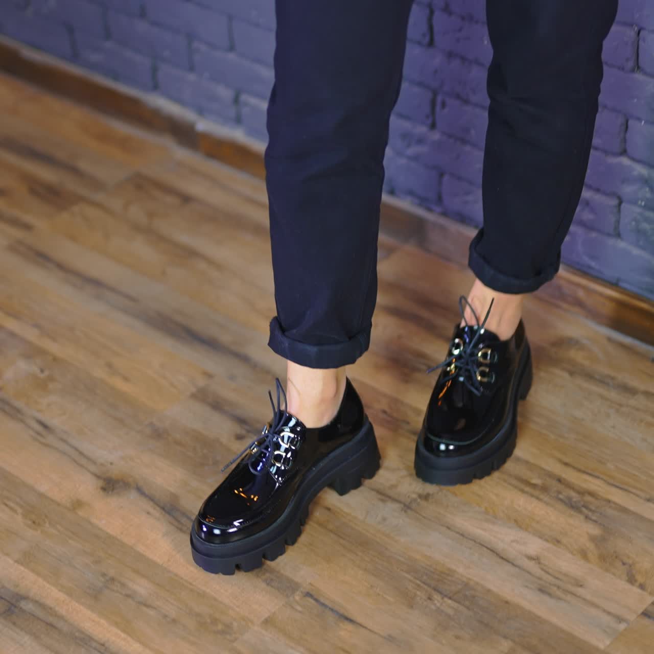 Woman wearing black massive shoes with laces and tractor soles. Lady stands leaned on the blue wall demonstrating fashionable footwear
