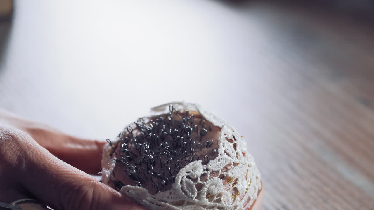 Female hand with beautiful nails taking pins from the white small pincushion. A lot of sewing pins in a sponge for dressmaker on the wooden table. Close-up.