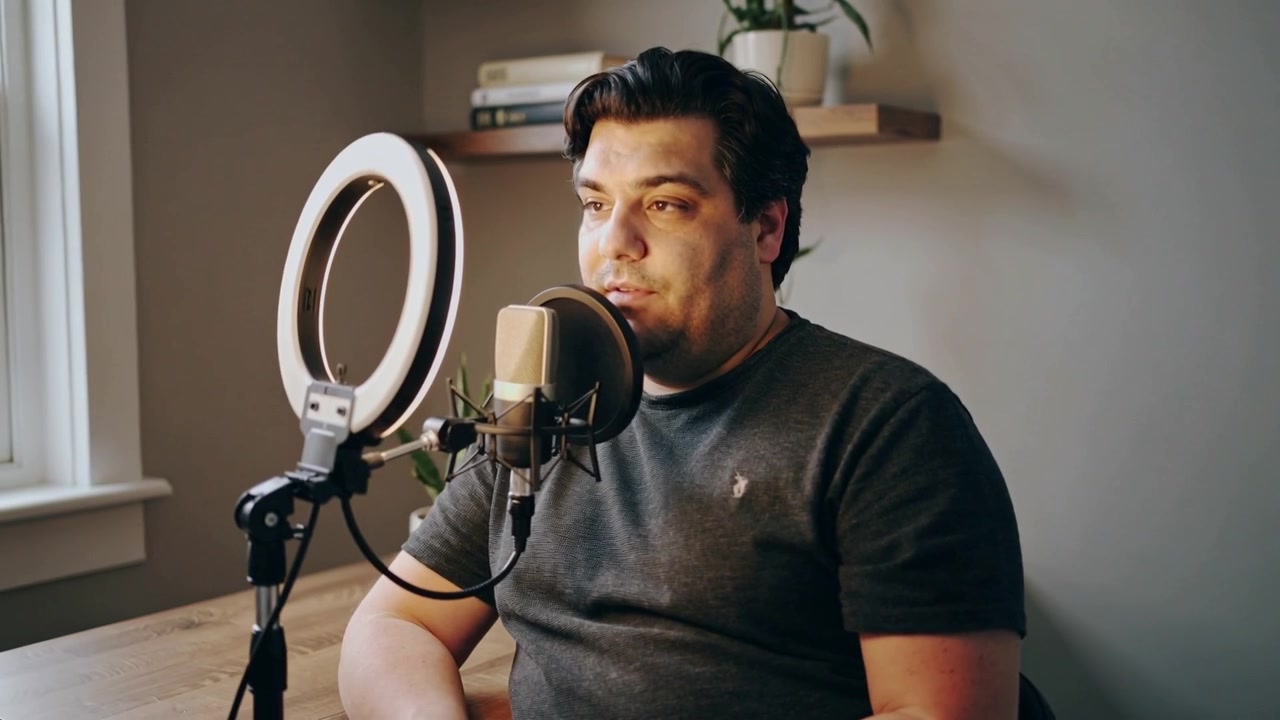 Recording starting host in dark tee speaking into mic by window, with ring light for podcast