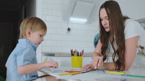 Mom-helps-sons-learn-to-draw-doing-homework-preschool-preparation-at-home-sitting-in-the-white-kitchen.