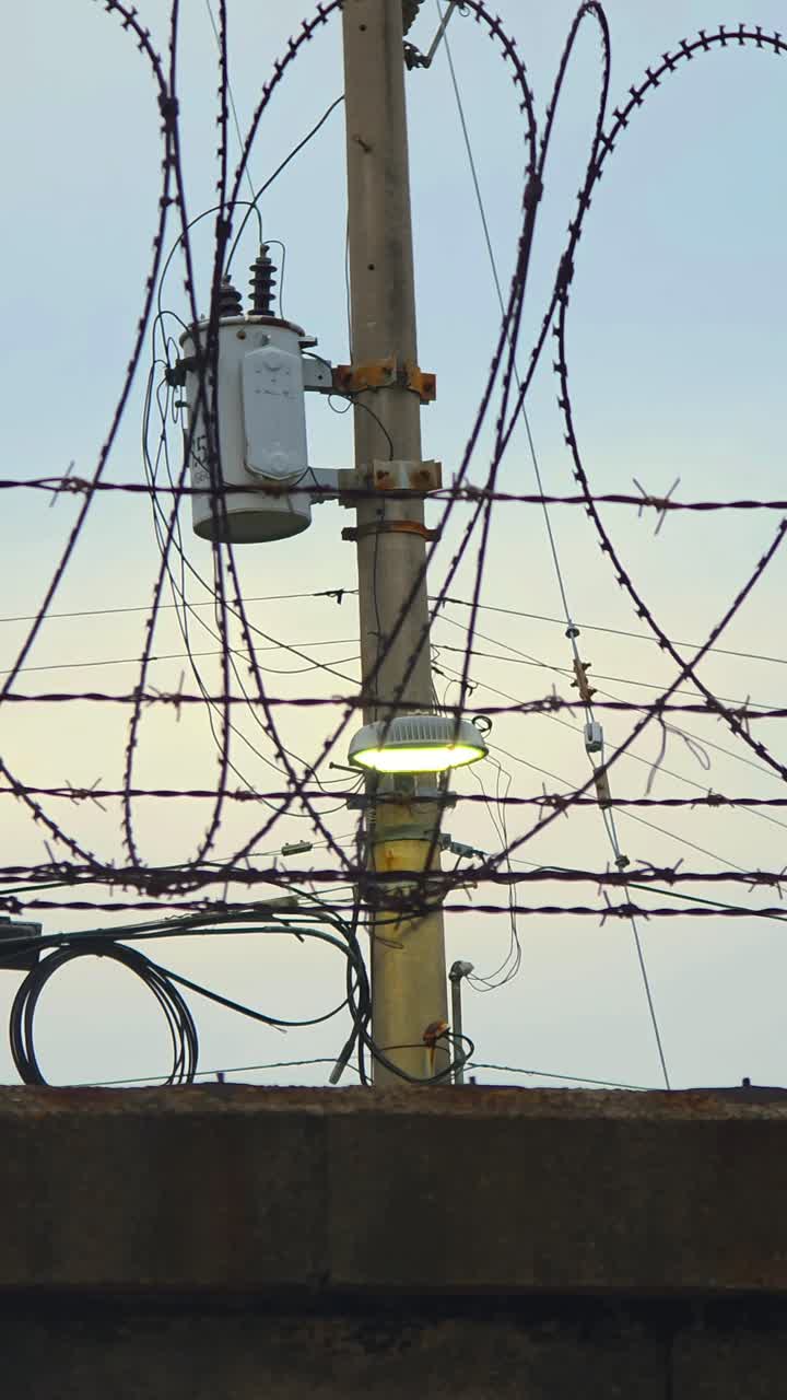 A trucking shot moves past a high-security fence with razor wire, focusing on a lit lamp and transformer on a utility pole, symbolizing a border, prison, division, or restricted area