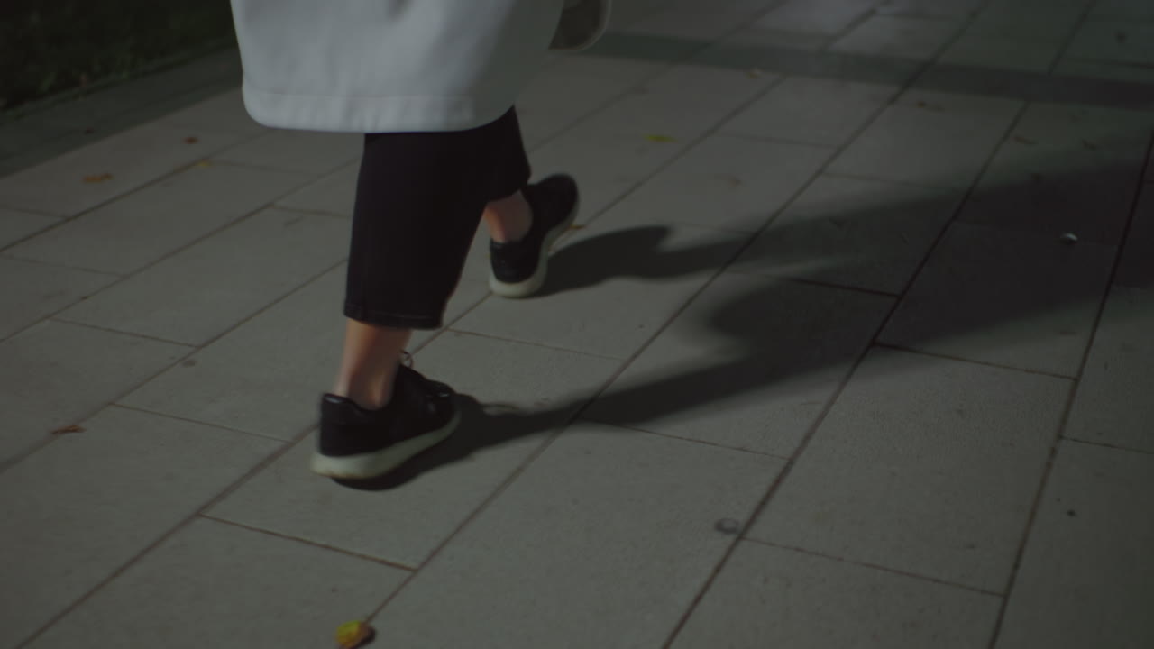 Leg view of lady wearing white coat and black trousers walking along tiled interlocked path at night, dimly lit with scattered foliage on ground, capturing peaceful ambiance of nighttime stroll