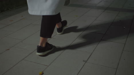 leg view of lady wearing white coat and black trousers walking along tiled interlocked path at night, dimly lit with scattered foliage on ground, capturing peaceful ambiance of nighttime stroll