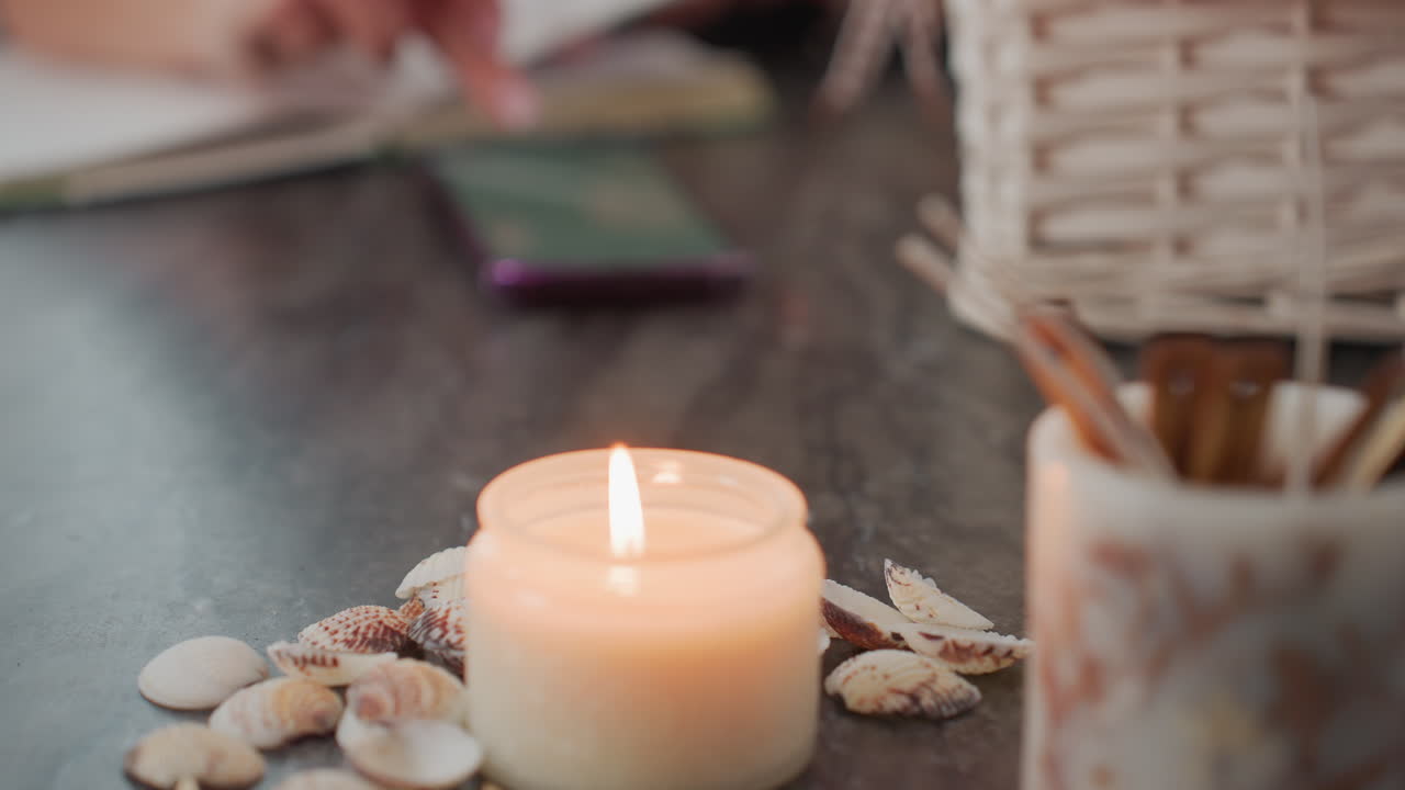 Cozy scene of candle lit up surrounded by seashells on dark table surface with blur view of person operating phone in background beside white basket in serene natural daylight through sheer curtains