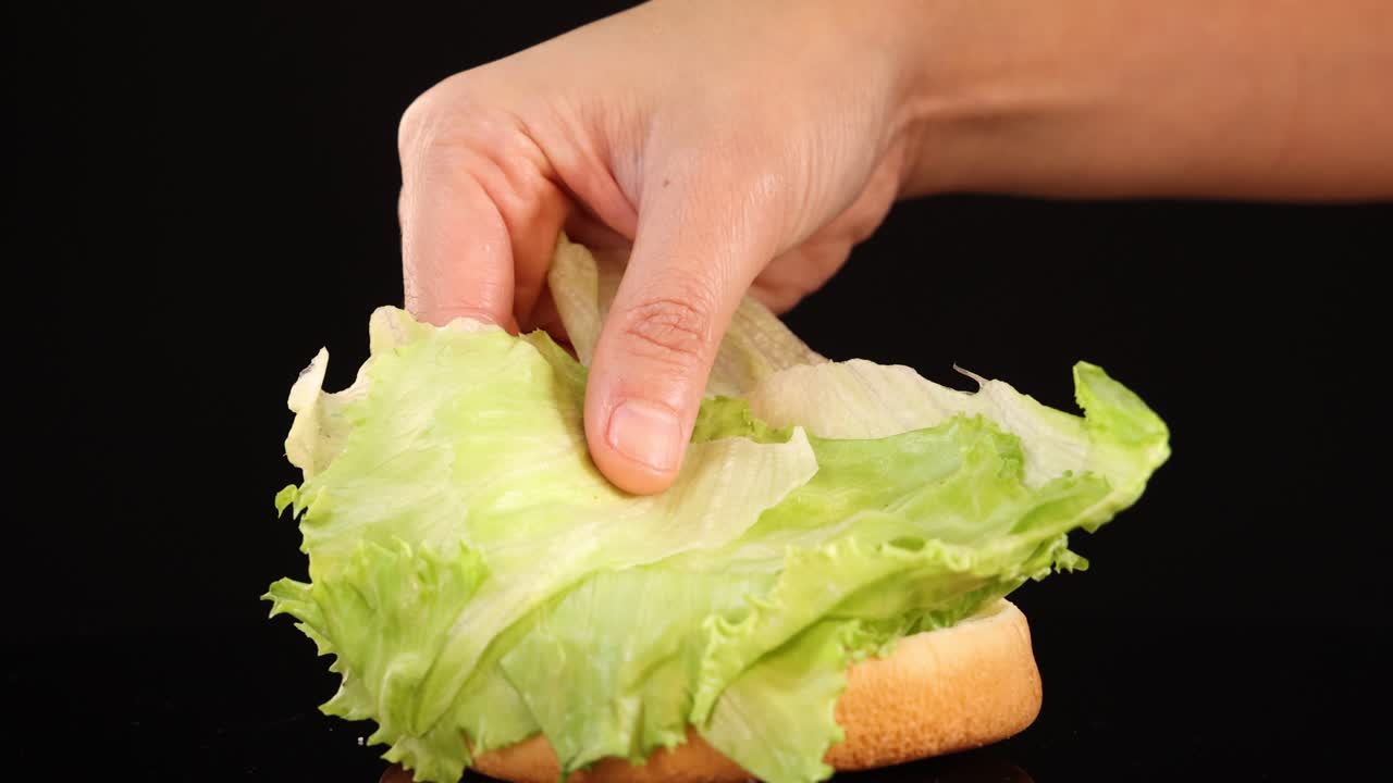A hand carefully arranges fresh iceberg lettuce onto a burger bun against a black background, using close-up shots and even studio lighting
