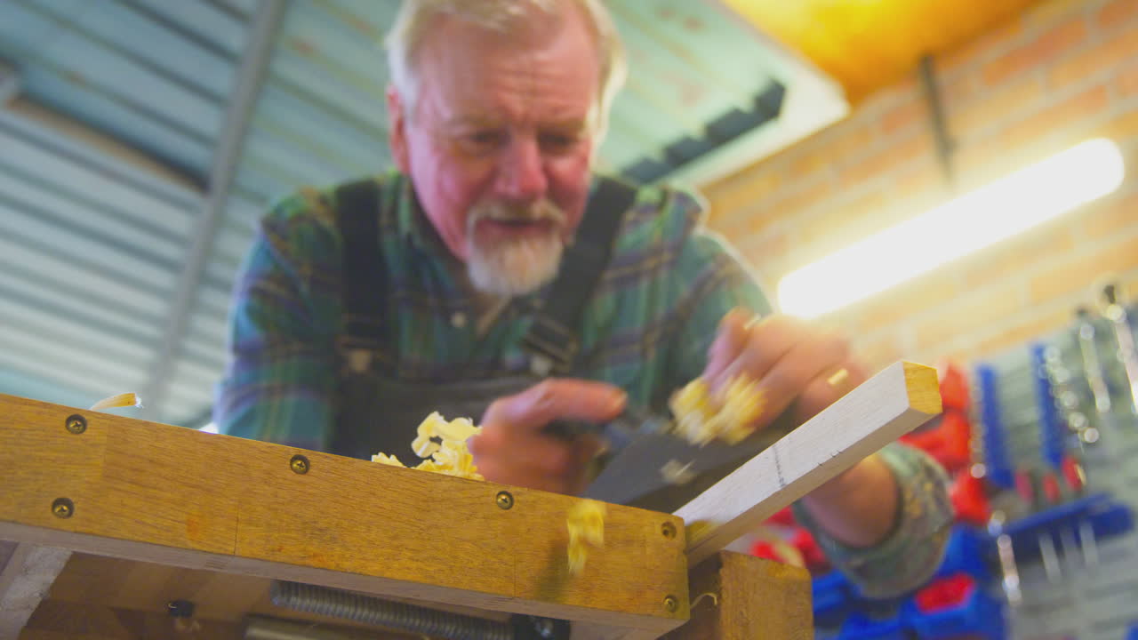 Close Up Of Senior Male Carpenter In Garage Workshop Planing Piece Of Wood