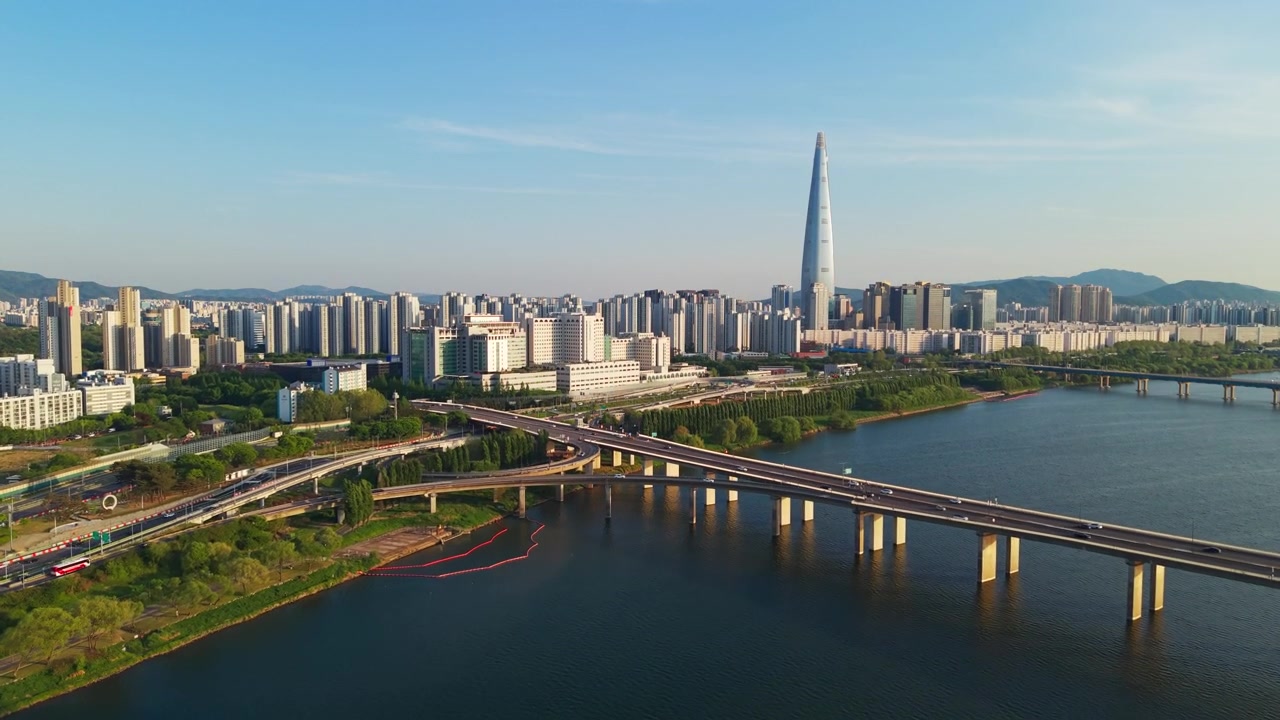 Ripresa aerea che rivela lo skyline della città di Seul, con l'iconico grattacielo Lotte World Tower e il traffico sul Ponte Jamsil che attraversa il fiume Han, sotto un cielo azzurro e limpido nel paesaggio urbano della Corea del Sud.