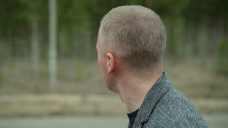 a close-up side view of an aged man with short hair, wearing a grey suit jacket and a black shirt, walking outdoors, the blurred background shows greenery