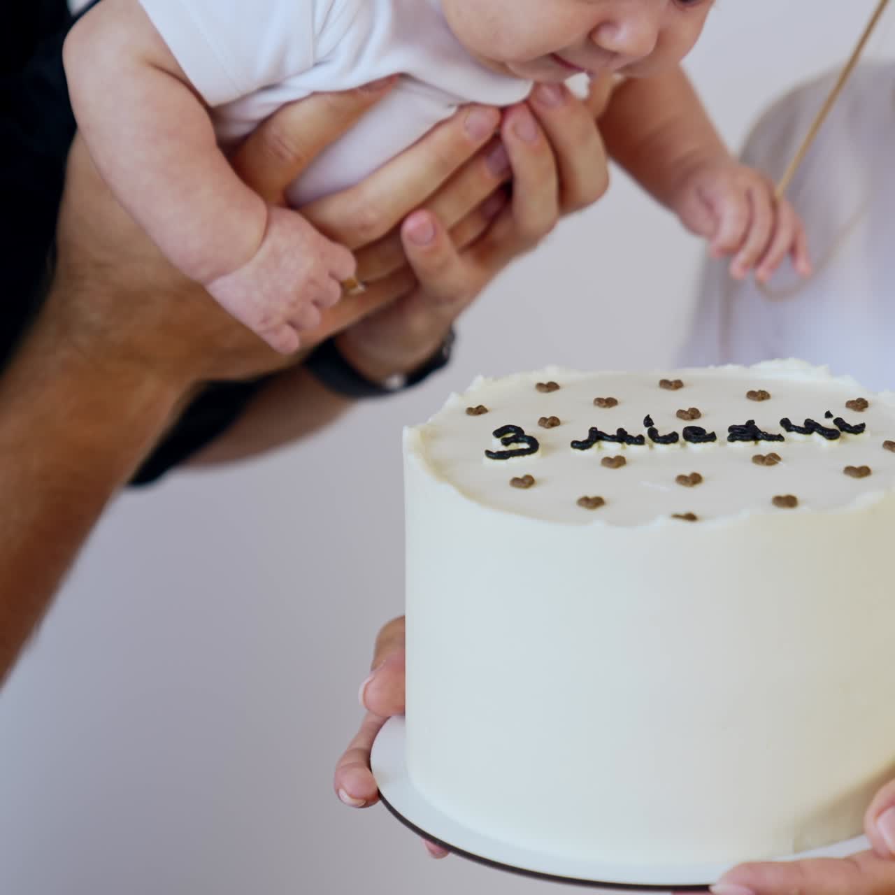 White cake with a black sign "three months" on in the female hands. Man holds a baby near the cake