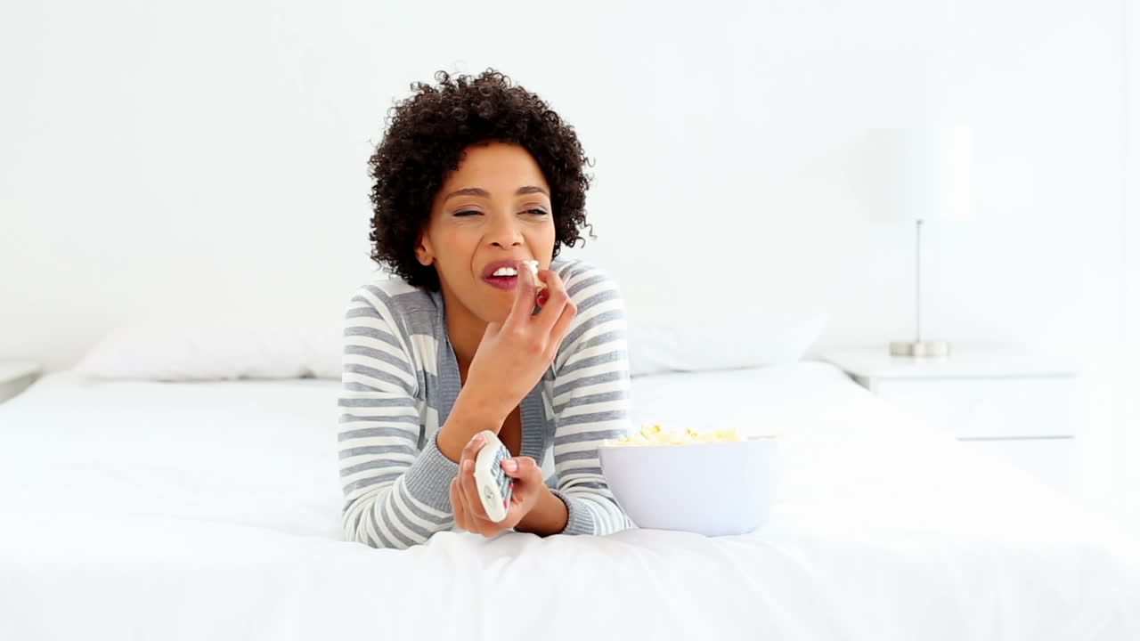 Sød, grinende kvinde ligger i sengen og ser tv