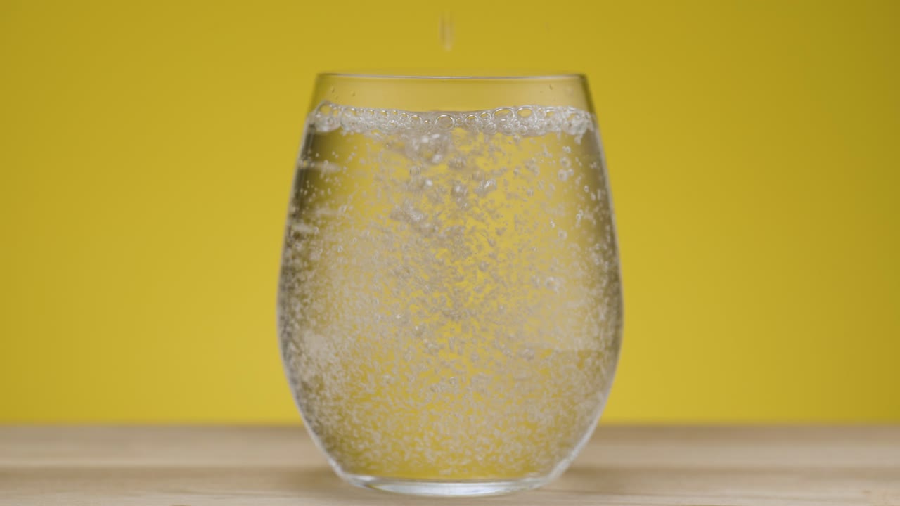vertiendo agua con gas en un vidrio transparente con fondo amarillo