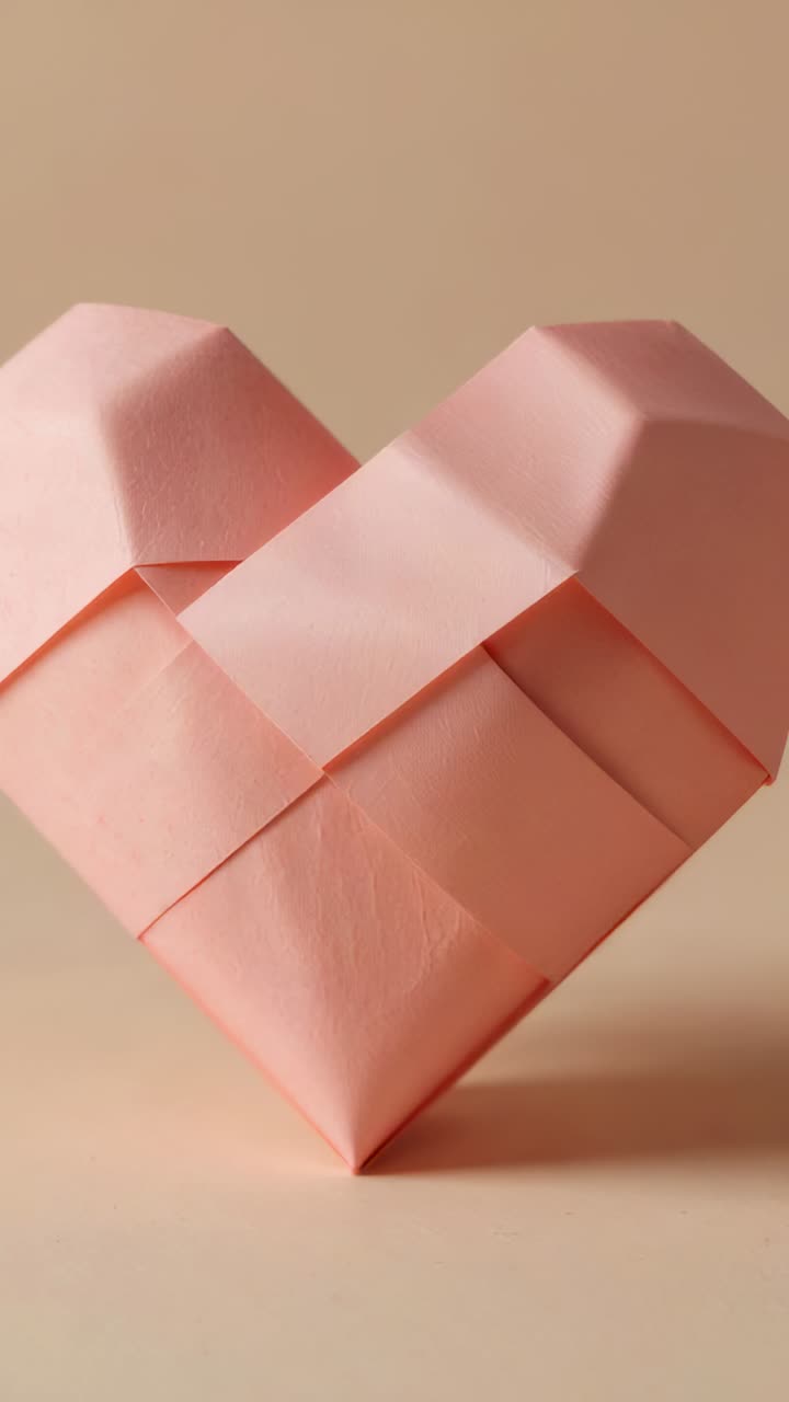 Vídeo vertical: reacción a un bokeh dorado, corazón de origami rosa inclinado sobre una mesa beige que revela los pliegues