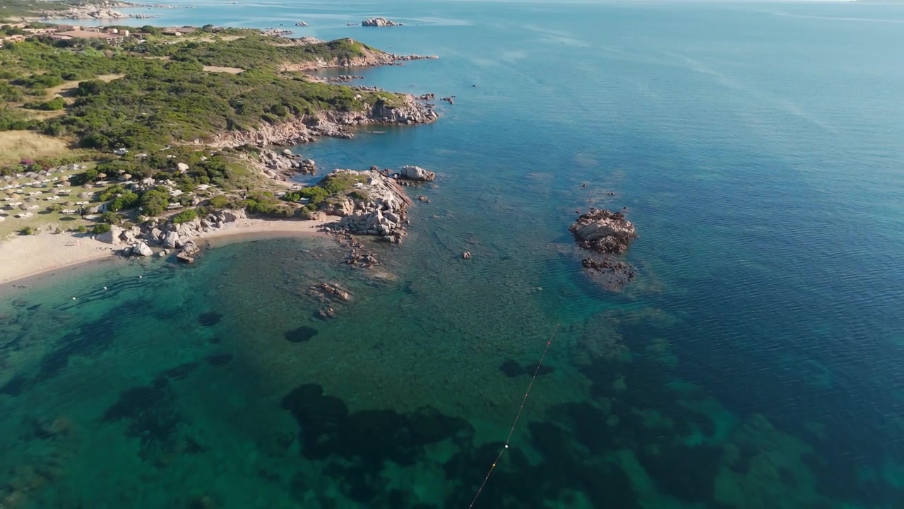Vista aerea che sorvola una costa rocciosa, acqua turchese limpida e una spiaggia sabbiosa.