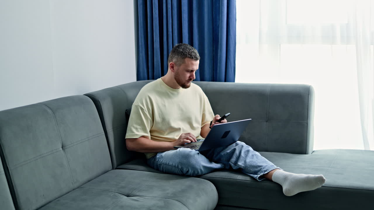 Man sits on gray couch using smartphone and laptop in bright indoor setting during the day