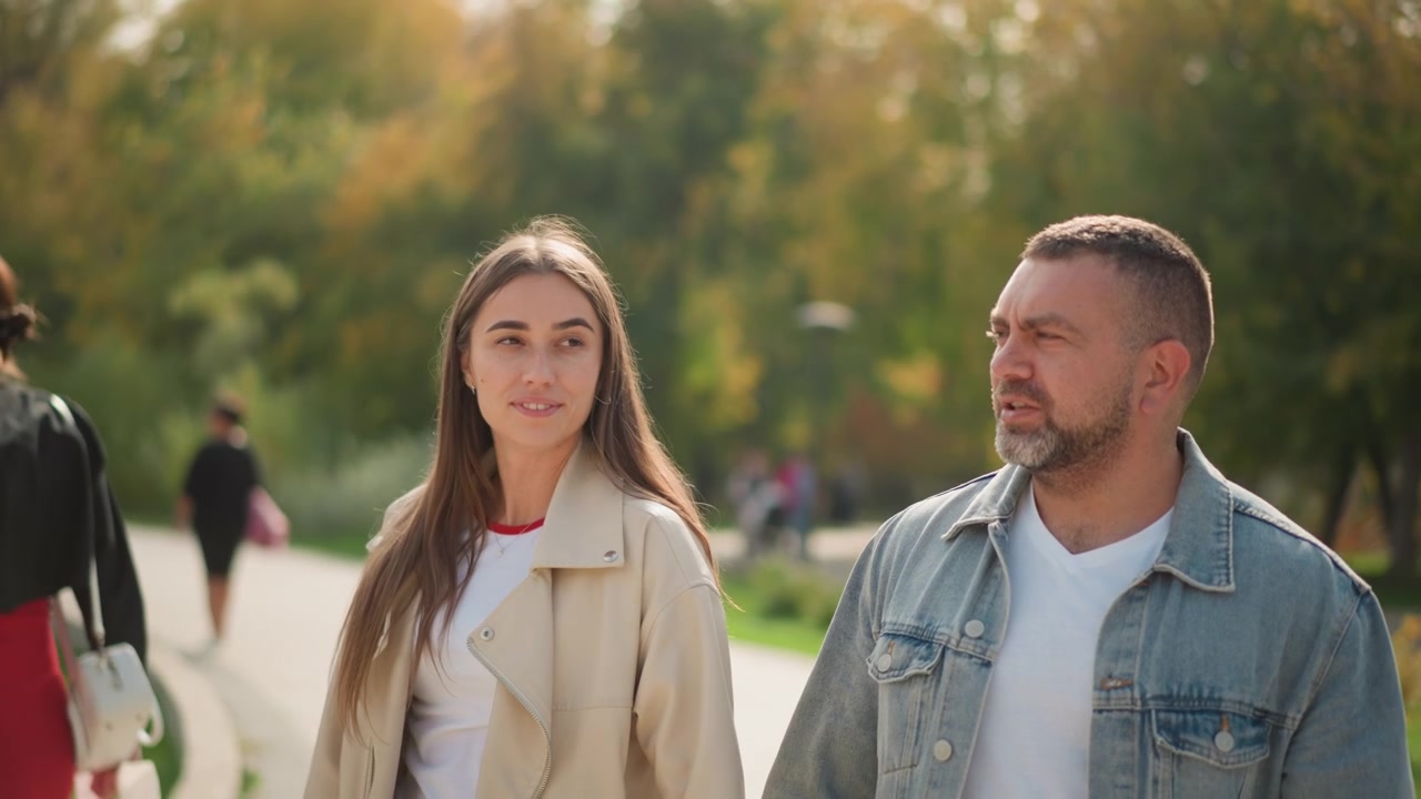 Primer plano de mujer joven y hombre caminando juntos en un parque, sonriendo. La mujer gesticula con la mano mientras ambos miran hacia adelante en un sendero rodeado de vegetación y un estanque. Iluminación suave.