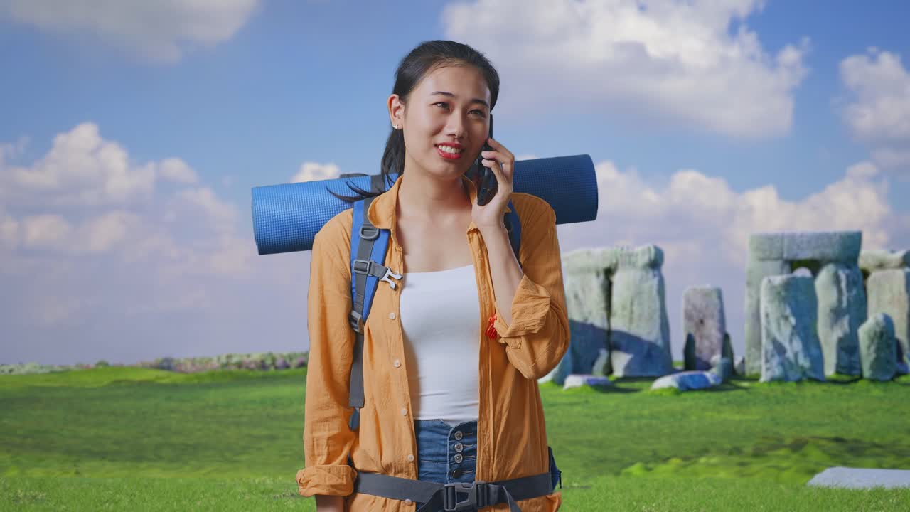 Asian Female Hiker With Mountaineering Backpack Smiling And Talking On Smartphone While Traveling In Stonehenge