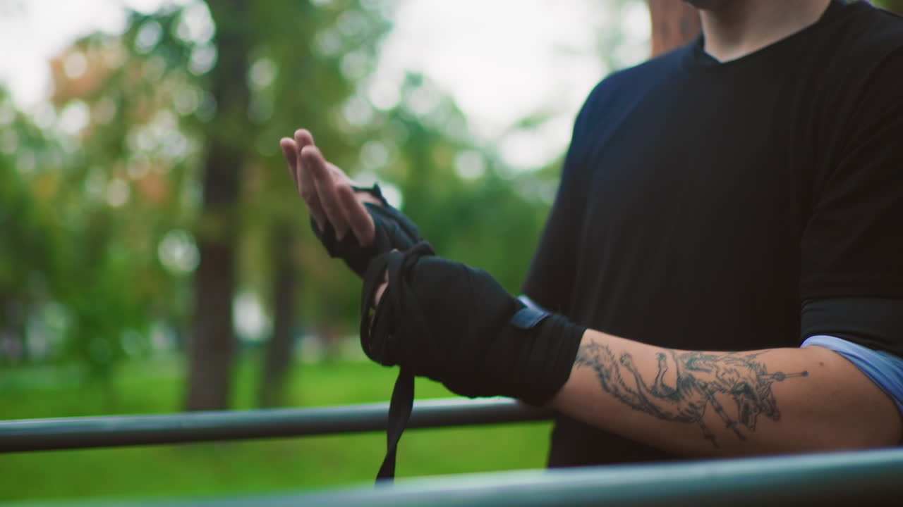 Ein weißer Mann sichert Handbandagen an einem Geländer, tätowierter Arm und schwarze Bänder, präzise Technik und Knotbewegung im Abendpark, methodisches Aufwärmen vor den Boxübungen, praktische Trainer-Demonstration